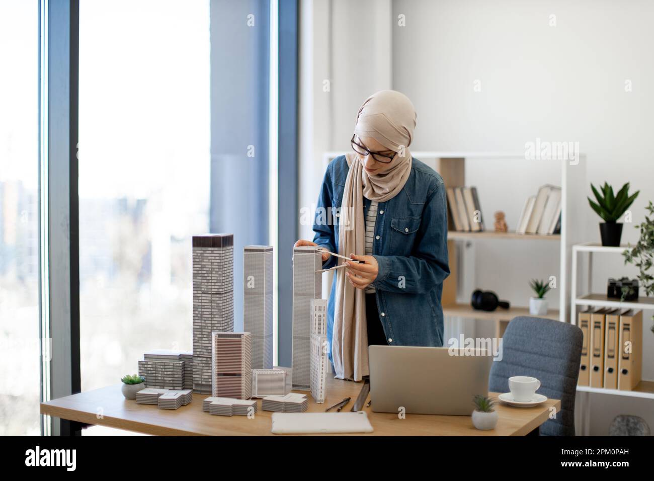 Arabian businesswoman in hijab and spectacles examining building model with compass while standing near writing desk with coffee and gadgets on it. Talanted architect using drawing tools at work. Stock Photo