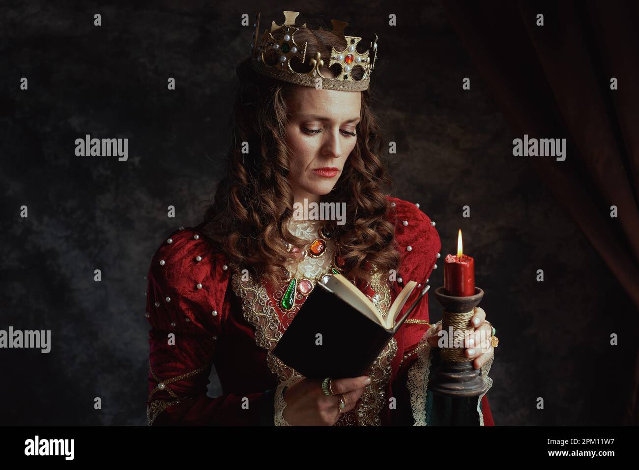 medieval queen in red dress with book, candle and crown on dark gray ...