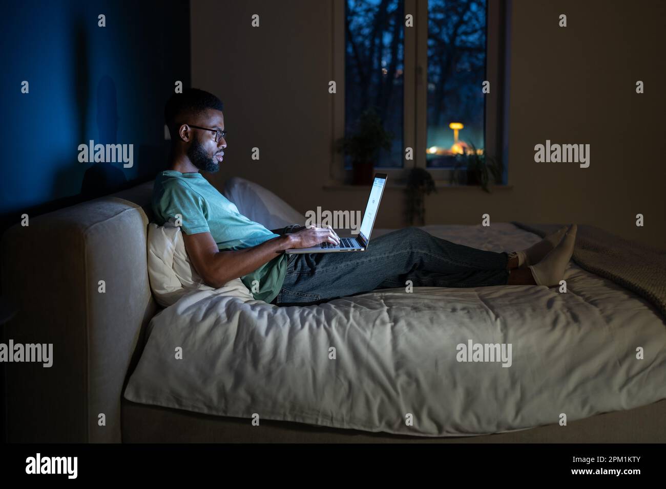 Black student guy lying in bed with laptop in evening, studying until late hours Stock Photo