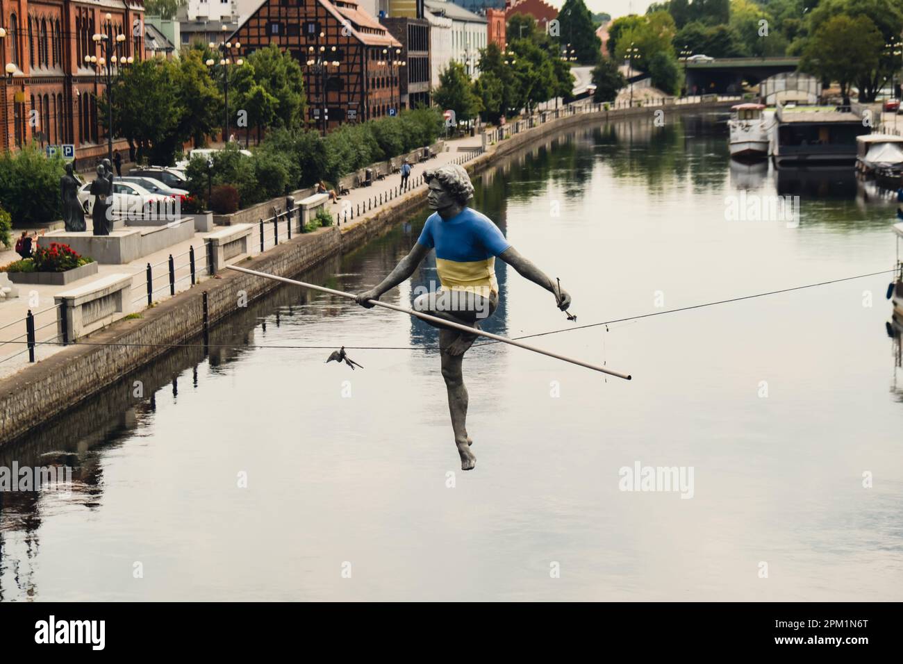 Bydgoszcz, Poland - August 2022 Brda river in Bydgoszcz, Man crossing a river sculpture , of a man balancing on a wire, old granary building, Kuyavian-Pomerania. Old town with reflection in Brda River. Old town view with famous statue in Ukrainian flag on the rope. Stock Photo
