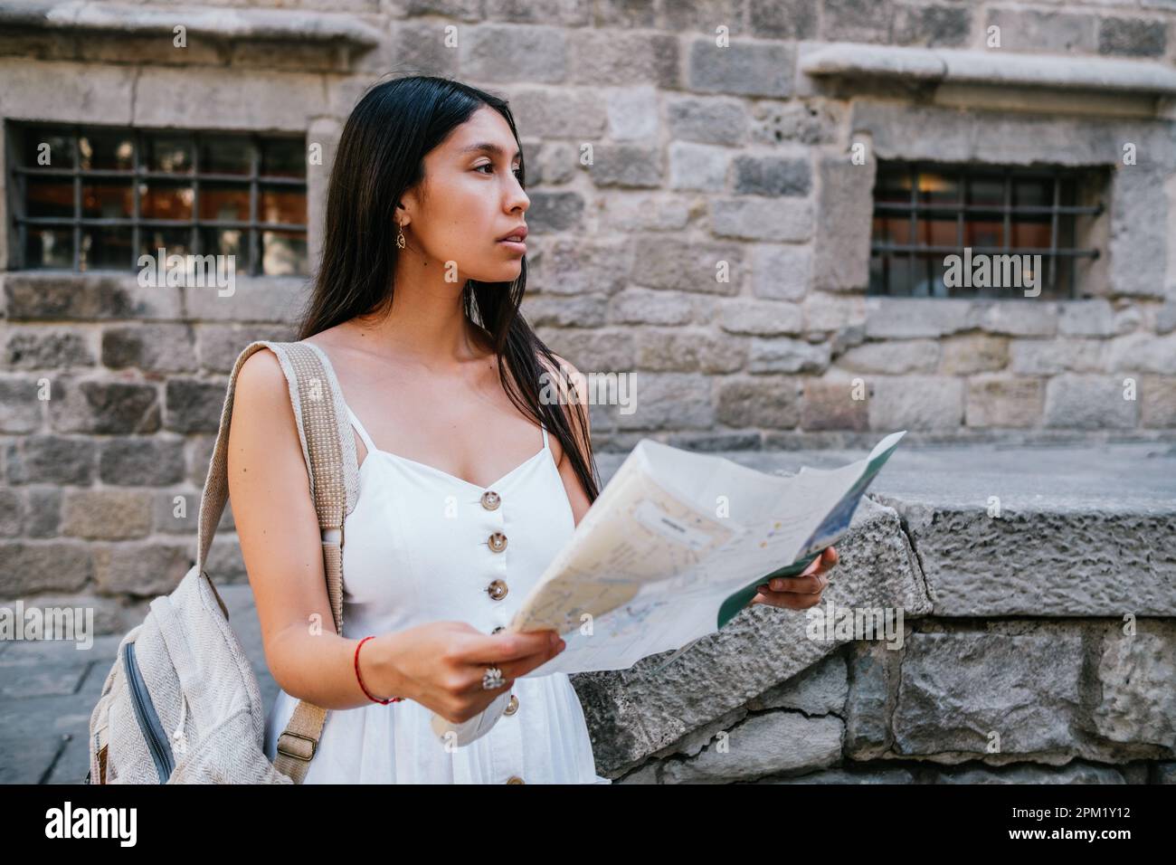 Hispanic woman in white dress with backpack and paper map looking away while standing near aged stone building on weekend day on street of Barcelona, Spain Stock Photo