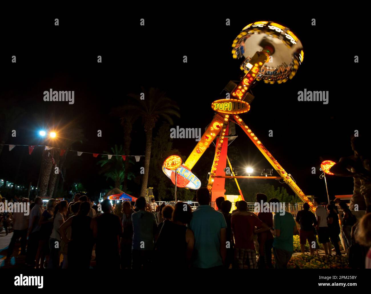 A crowd of people watch thrill seekers spin in mid air on the Jupiter ride at the amusement park set up at Antalya in Turkiye. Stock Photo