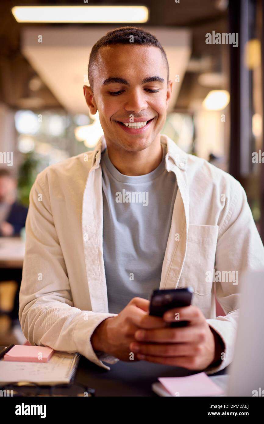 Young Businessman Working On Laptop At Desk In Office Checking Messages On Mobile Phone Stock Photo