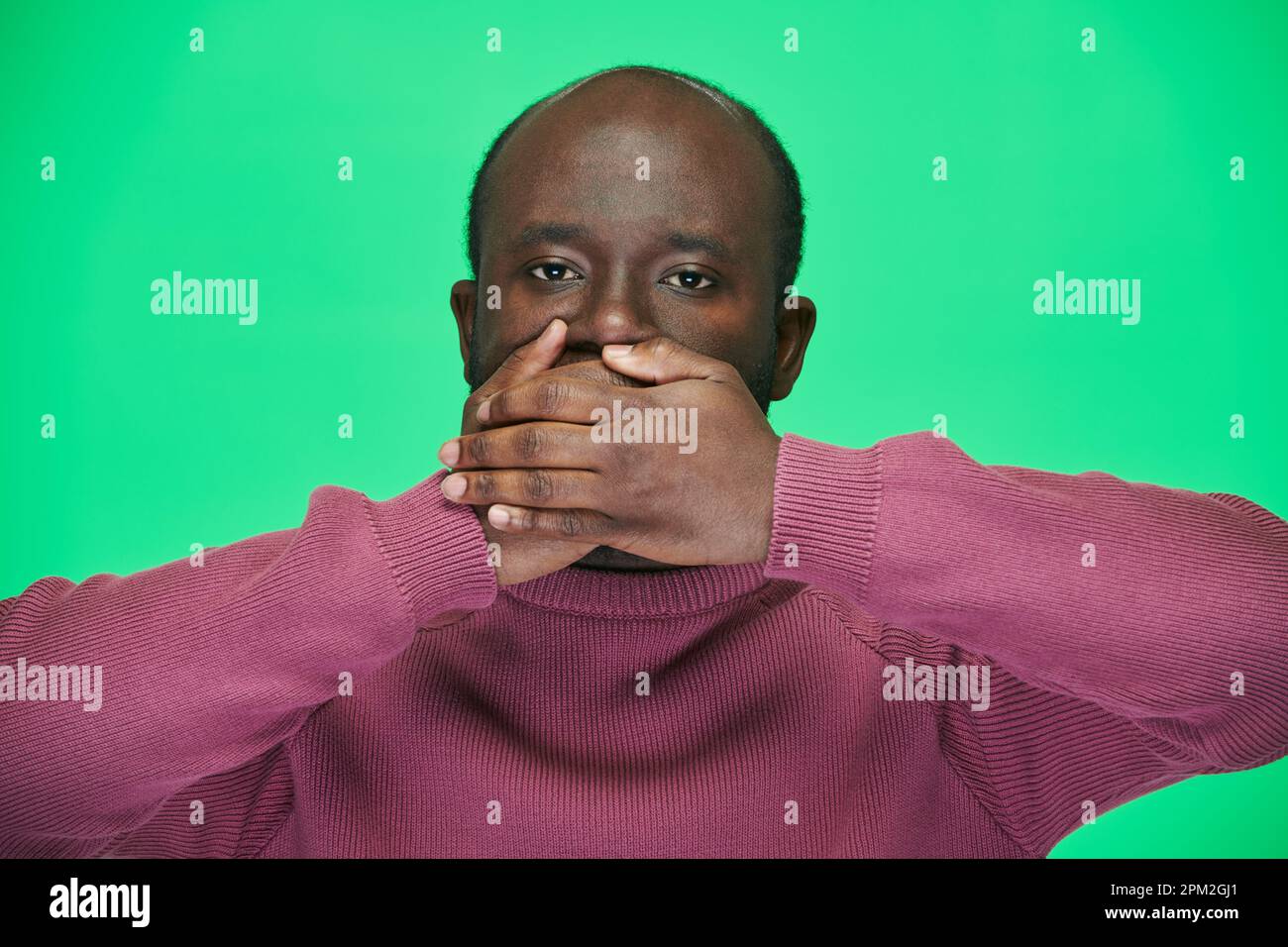 Portrait of African American man looking at camera closing his mouth with hands against green background Stock Photo