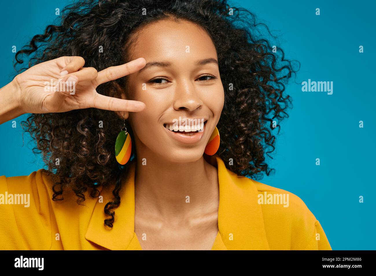 Studio portrait of joyful curly African American woman wearing hipster clothing showing v-sign near her face on blue background Stock Photo