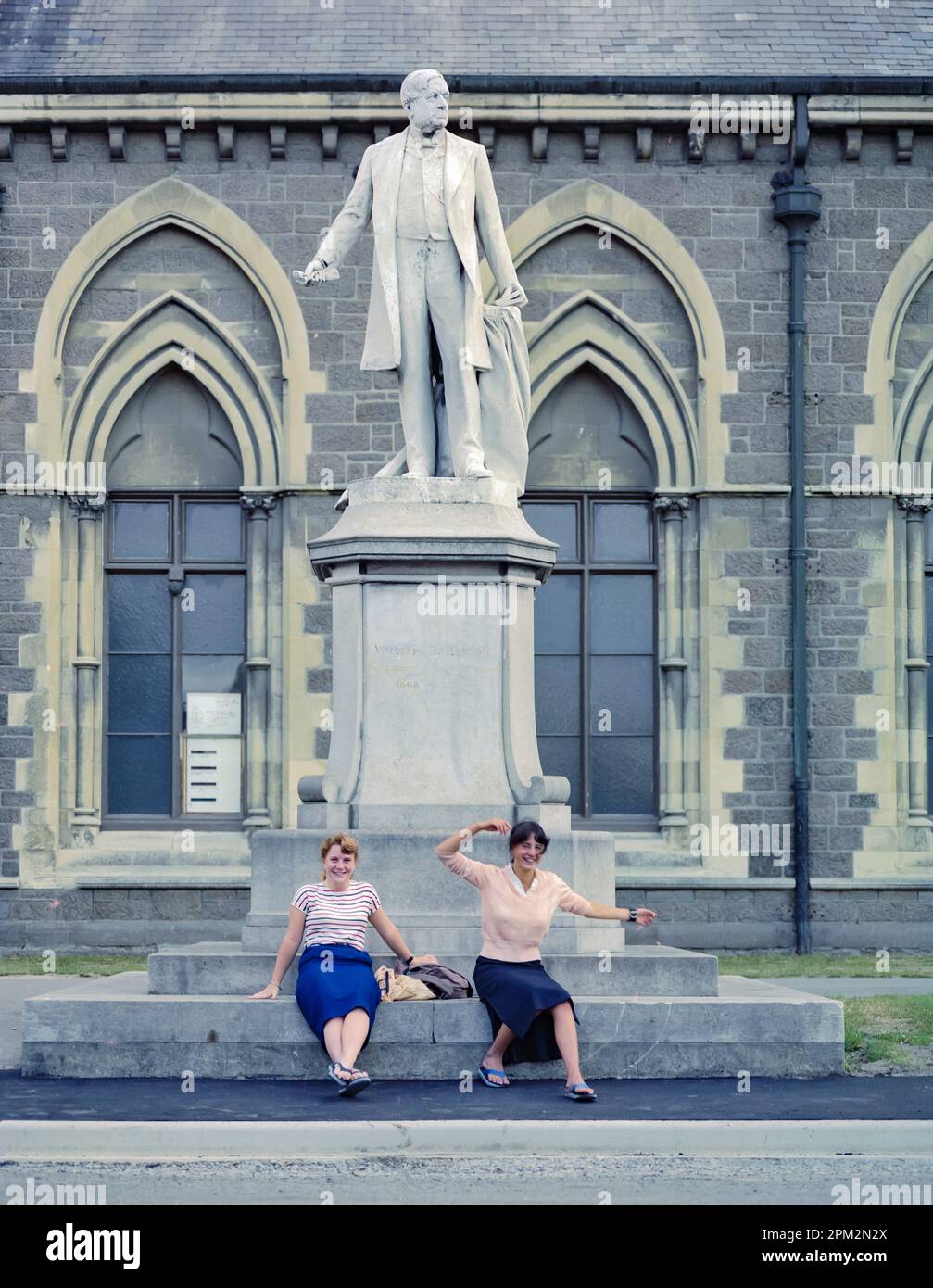 A 1981 historic image of two young women waving and play-acting at the base of the William Rolleston statue outside the Canterbury Museum in the city of Christchurch on the south island of New Zealand. In the 2011 Christchurch earthquake, the statue toppled off its plinth and was damaged but it was repaired and reinstated in 2016. Stock Photo