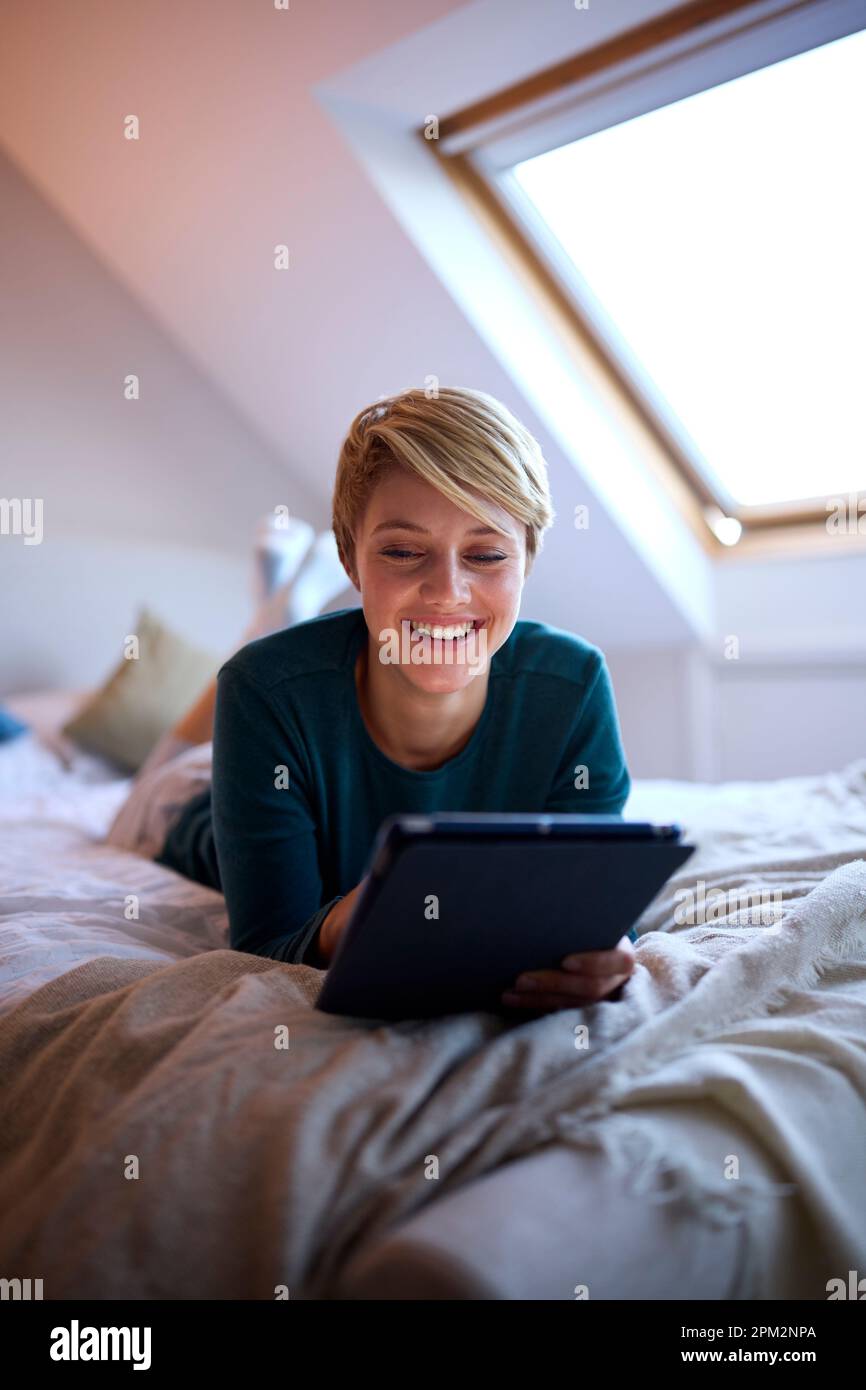 Young Woman Lying On Bed Wearing Pyjamas At Home Looking At Digital Tablet Stock Photo