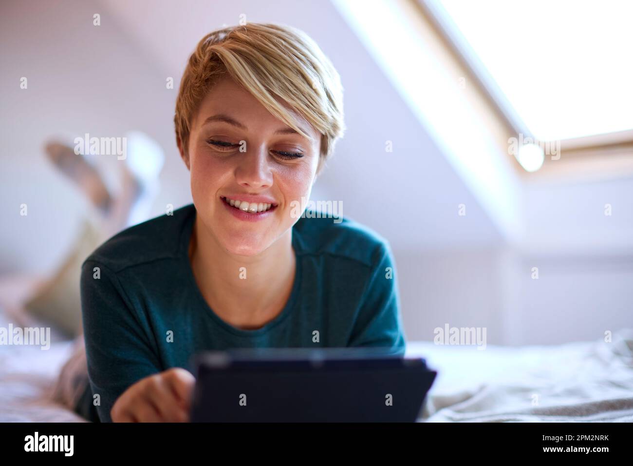 Young Smiling Woman Lying On Bed Wearing Pyjamas At Home Looking At Digital Tablet Stock Photo