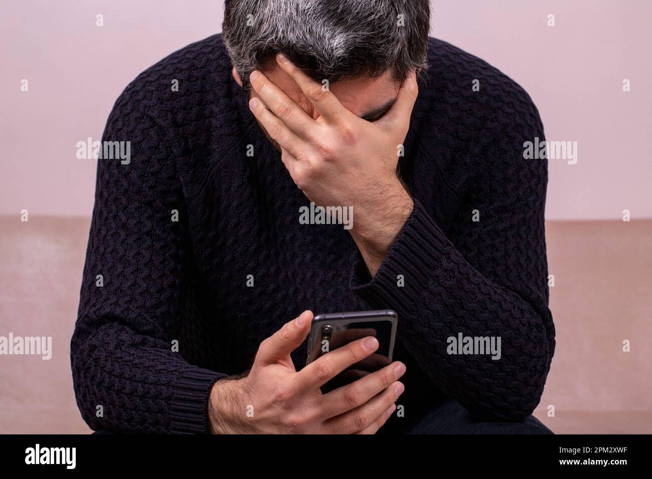 Middle-aged man receiving sad news on the phone. Concerned man reading news online looking at phone screen, Caucasian sitting on sofa at home, serious Stock Photo