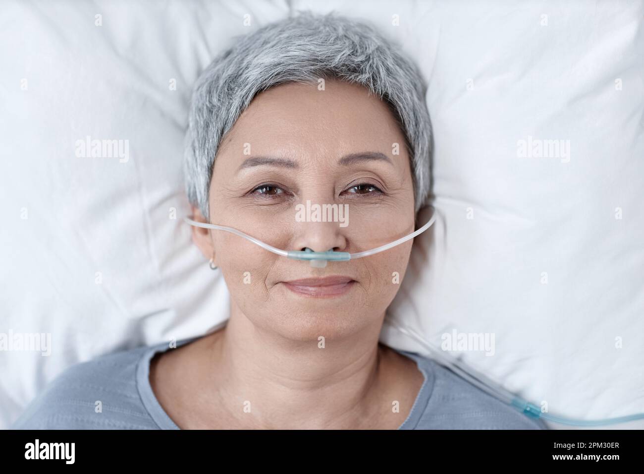 High angle view of senior woman in oxygen tube in her nose looking at camera while lying on bed in ward Stock Photo