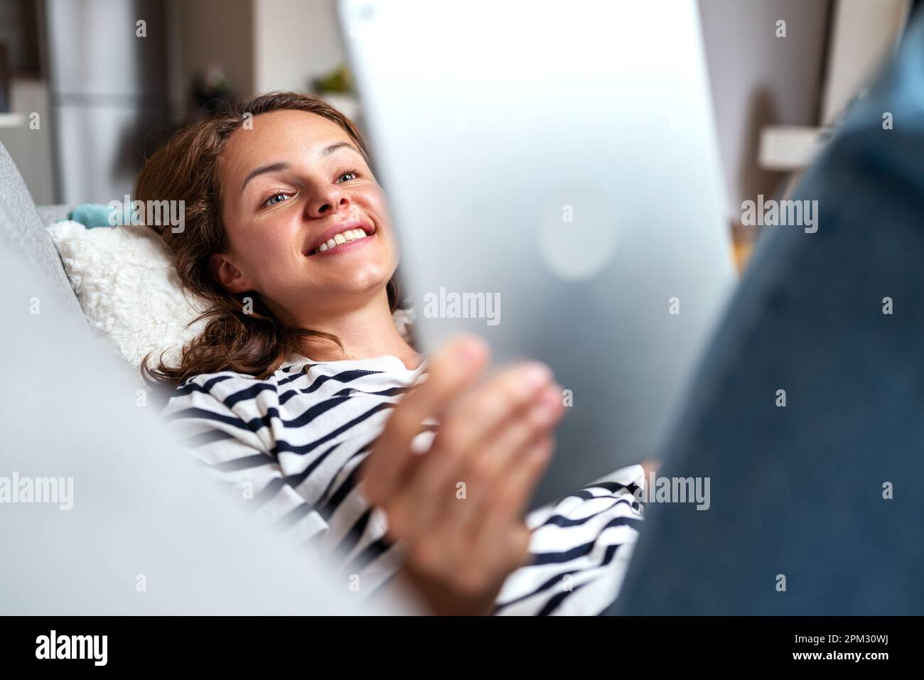 Woman smiling and using her digital tablet at home lying down on sofa. Stock Photo