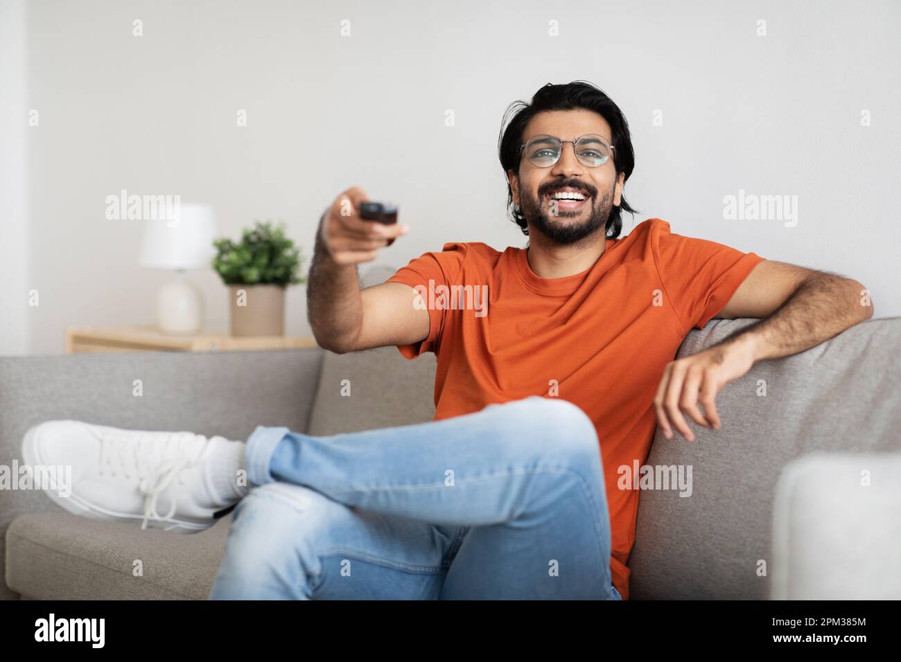 Happy young indian male in glasses sits on sofa with remote control, watching TV, enjoy free time Stock Photo