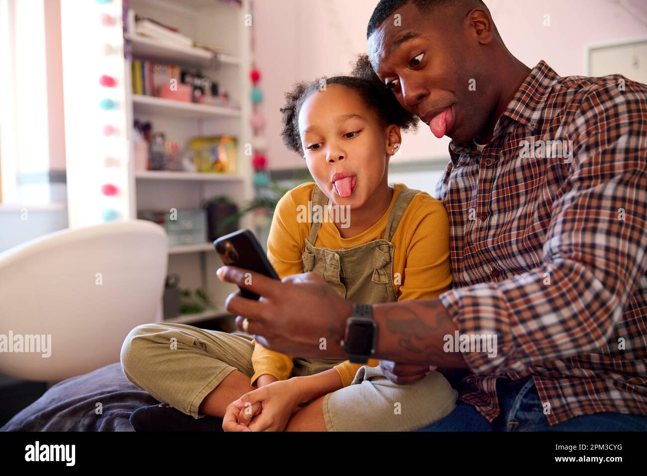 Father And Daughter At Home In Girl's Bedroom Posing For Selfie On Mobile Phone Pulling Faces Stock Photo
