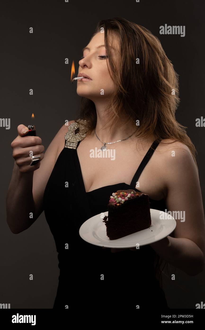 lighting a cigarette with a lighter and holding a piece of chocolate cake, young woman in a dress in studio, addictions and sweet food, dessert Stock Photo