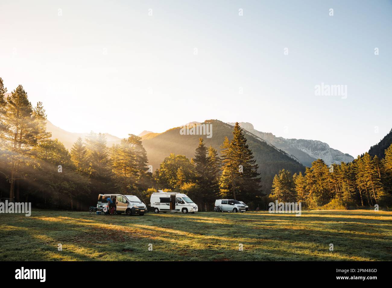 Camper vans in a valley with amazing landscape views of forest, mountains and sun rays during sunrise. Van road trip holiday and outdoor summer advent Stock Photo