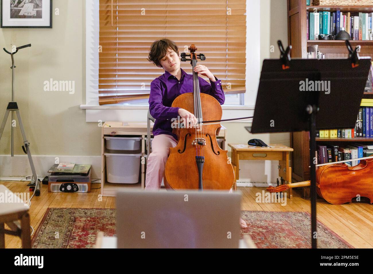 A young passionate boy plays cello in front of computer Stock Photo