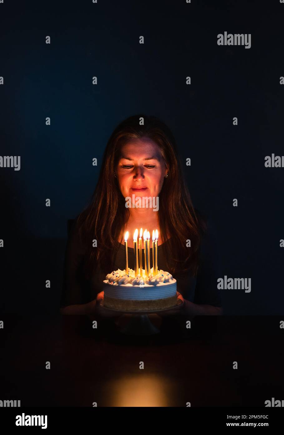 Attractive woman looking at candles on birthday cake in dark room. Stock Photo