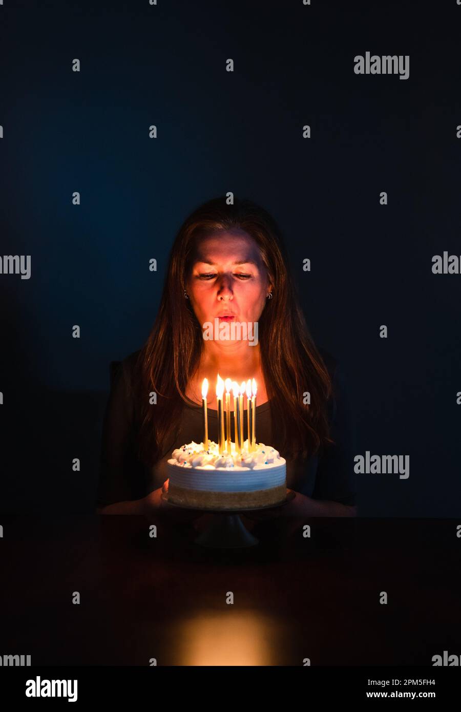 Attractive woman blowing out candles on birthday cake in dark room. Stock Photo