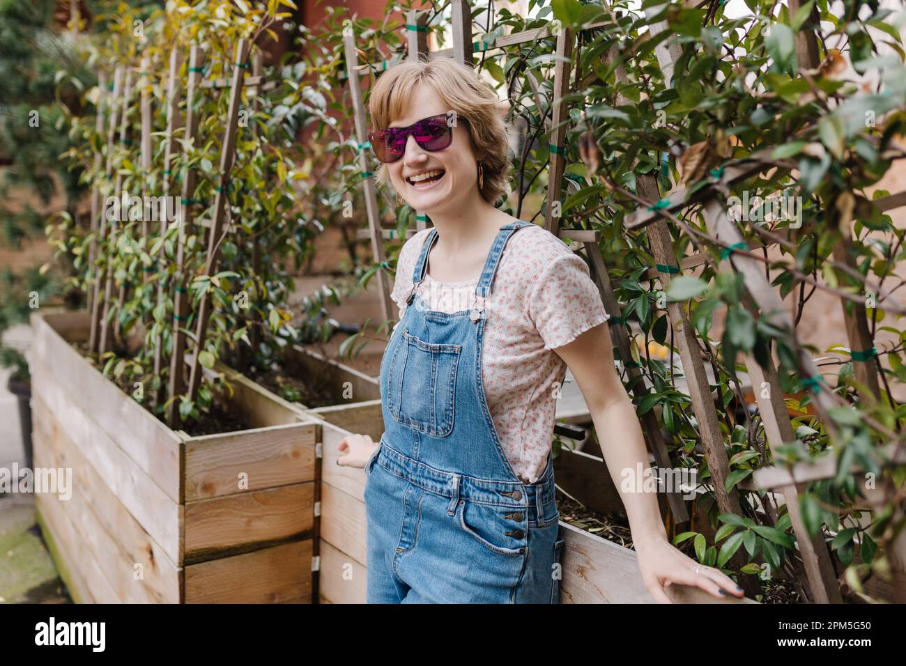 Fashionable woman smiling with sunglasses leaning against plants Stock Photo