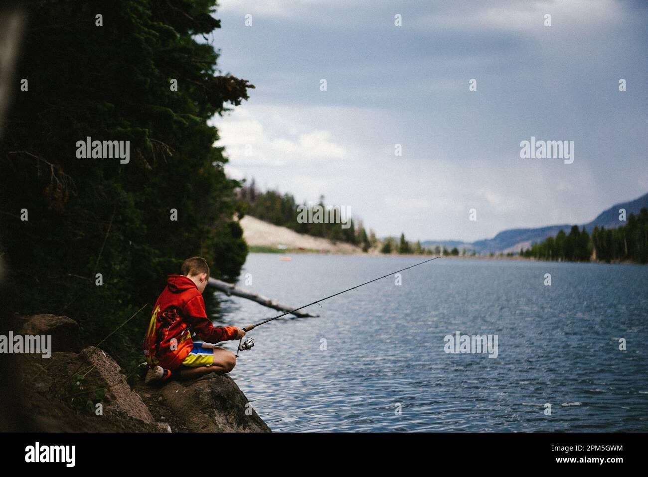 Boy fishing in a lake in mountains with forest around him Stock Photo