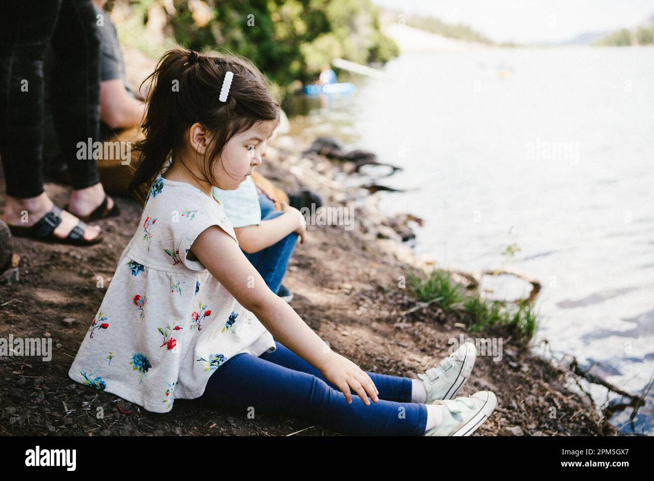 Little girl wearing a dress and siting on lake shore with family Stock Photo