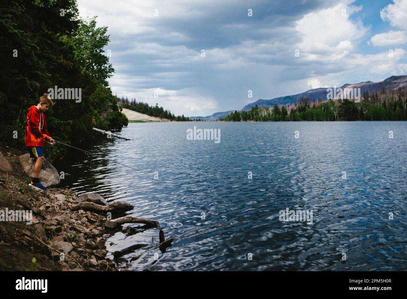 Boy fishing on lake with forest, mountains in outdoor adventure Stock Photo