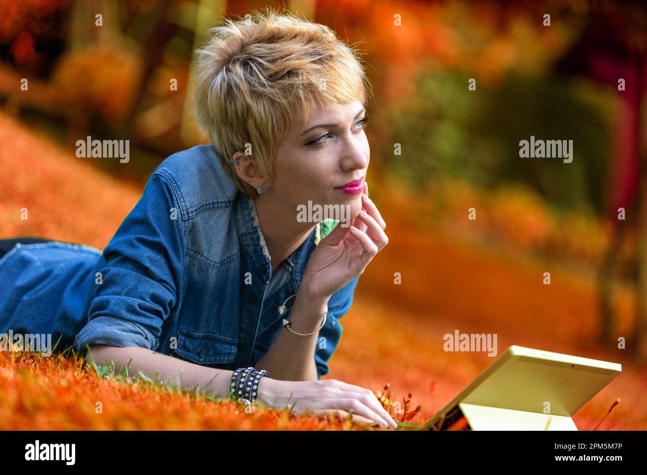 Colorful portrait of a young woman lying face-down on the grass while using a laptop-tablet combo. In the midst of nature, well-off, she can afford to Stock Photo
