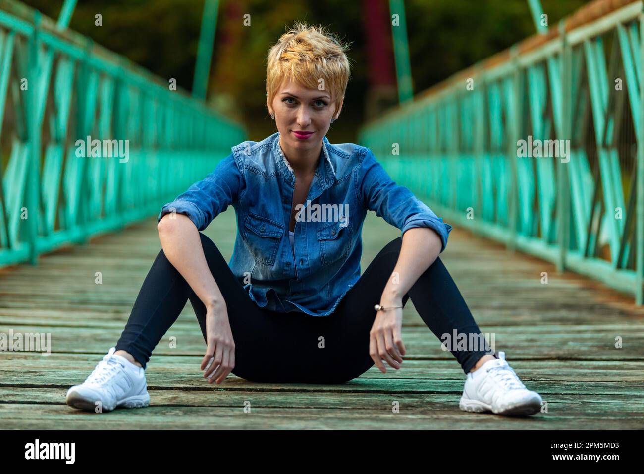 Bossy pose, fighter style, for this beautiful blonde-haired woman sitting confidently in the middle of the bridge in the mountains, suspended over the Stock Photo
