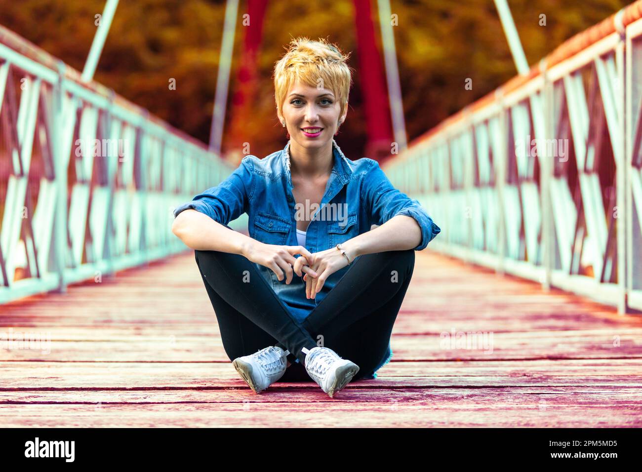 Bossy pose, fighter style, for this beautiful blonde-haired woman sitting confidently in the middle of the bridge in the mountains, suspended over the Stock Photo