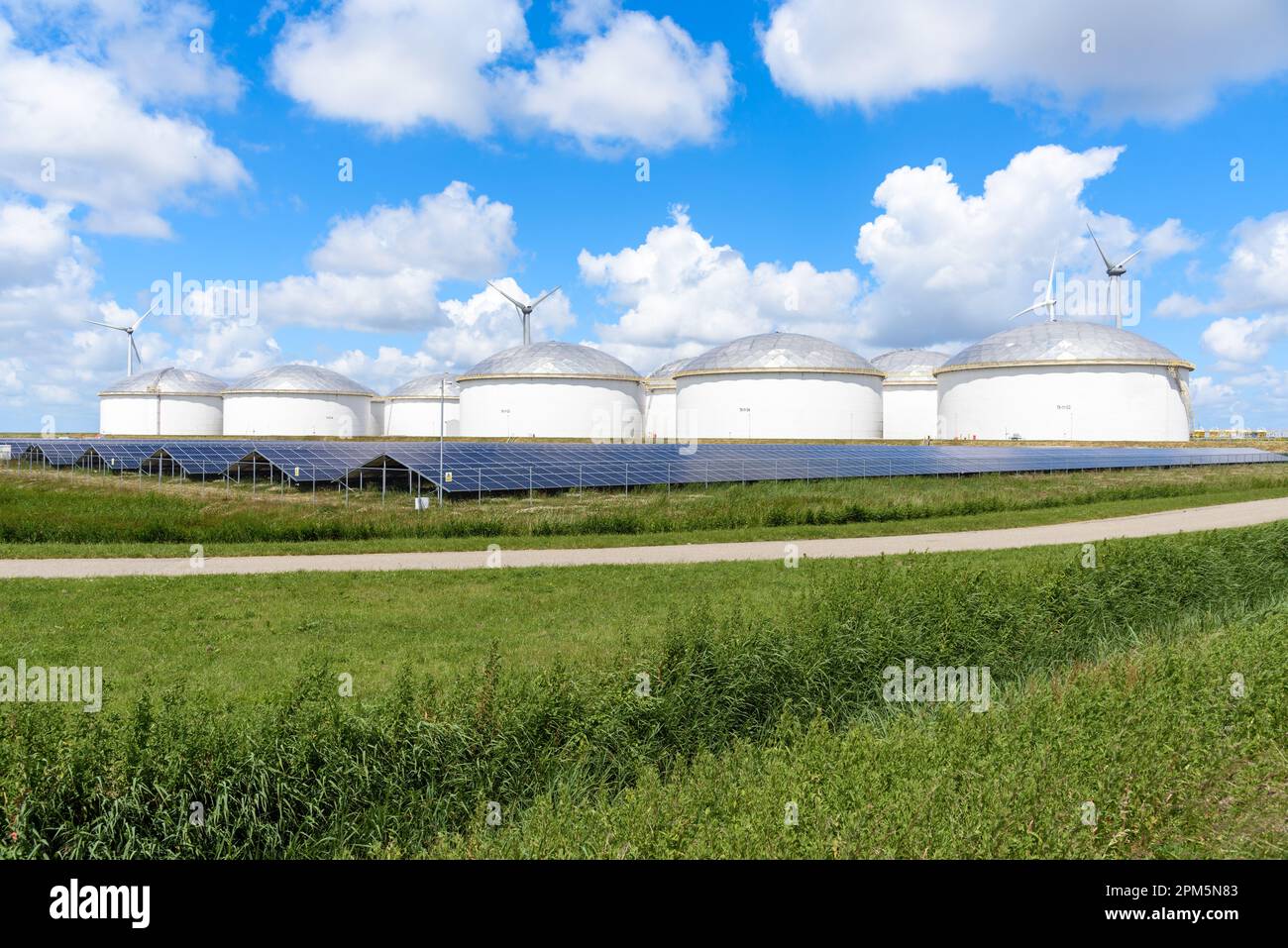 Row of solar panels in front of a group of fuel storage tanks in a business park on a sunny summer day. Wind turbines are visible in background. Stock Photo