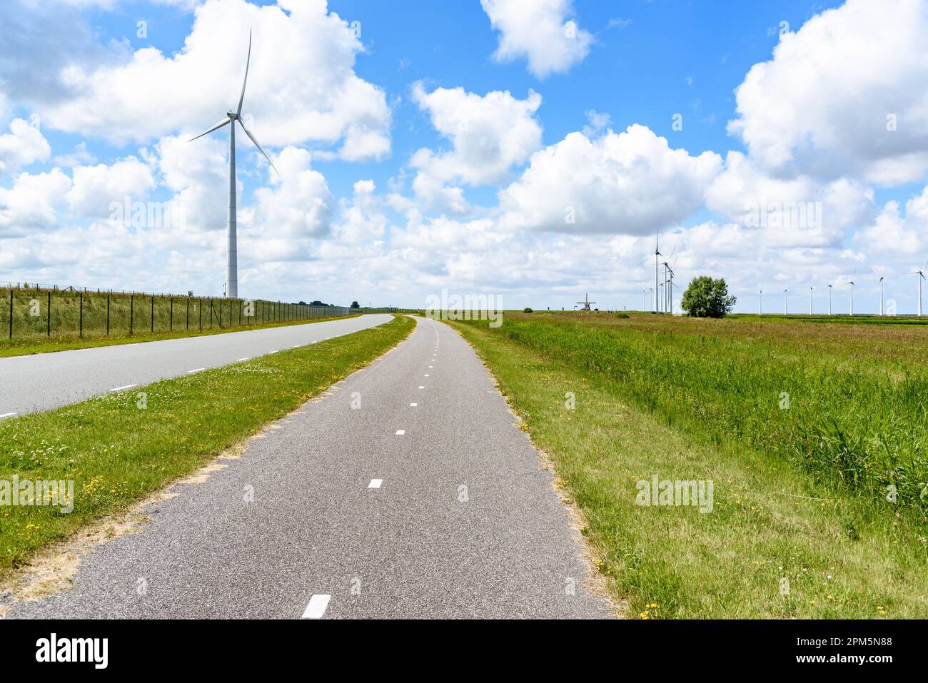 Empty bicycle path overlooked by a tall wind turbine in the countryside of Netherlands on a sunny summer day. A wind farm is visible in distance. Stock Photo