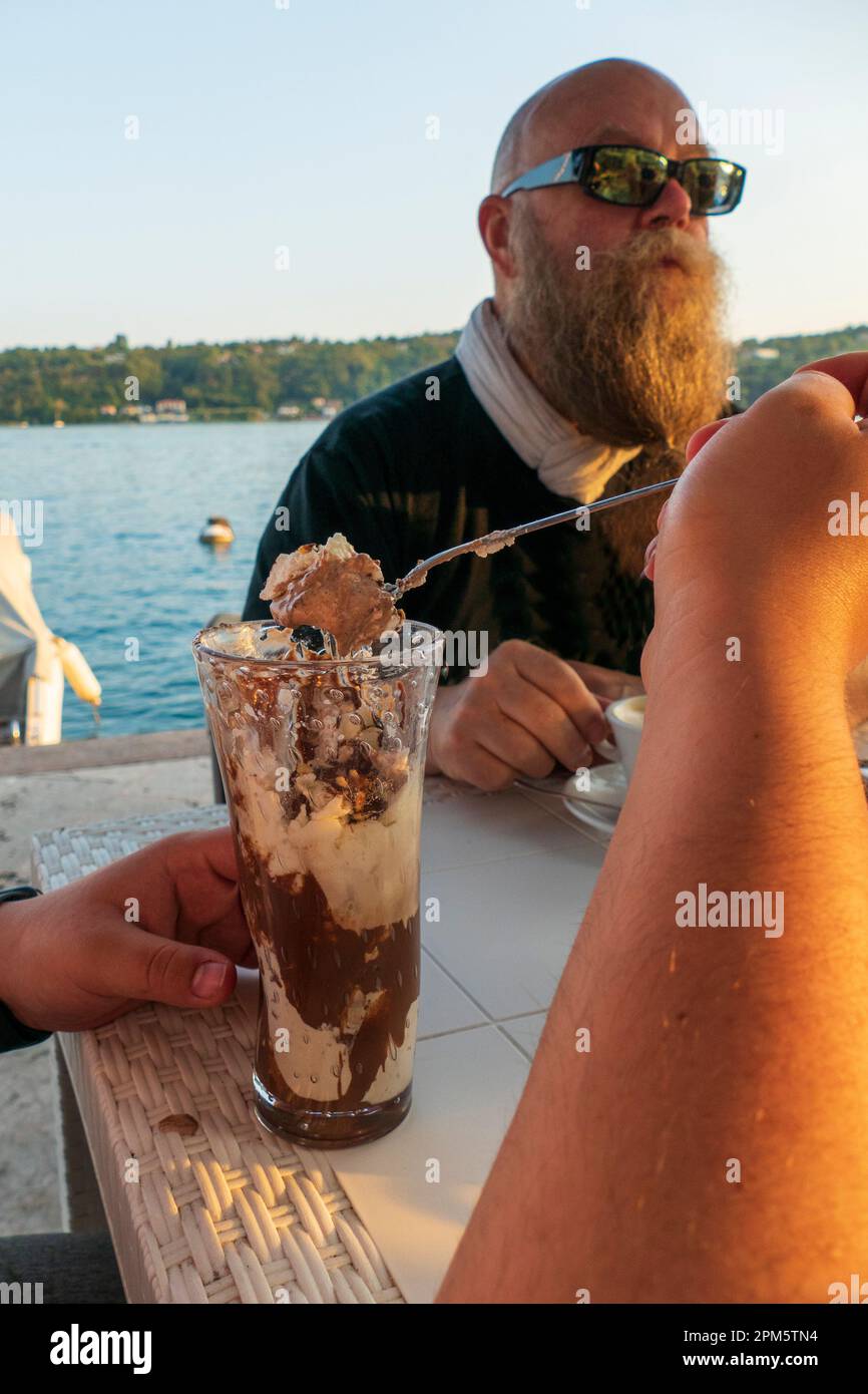 Eine Person isst Eiscreme, im Hintergrund ein Mann mit langem Bart an einem See Stock Photo