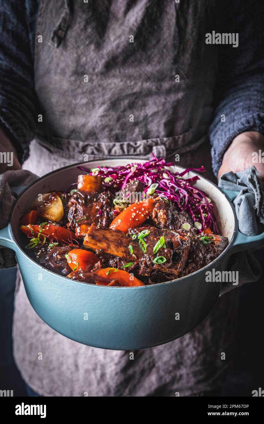 Man in gray apron holding Dutch Oven with Braised Short Ribs, carrots ...
