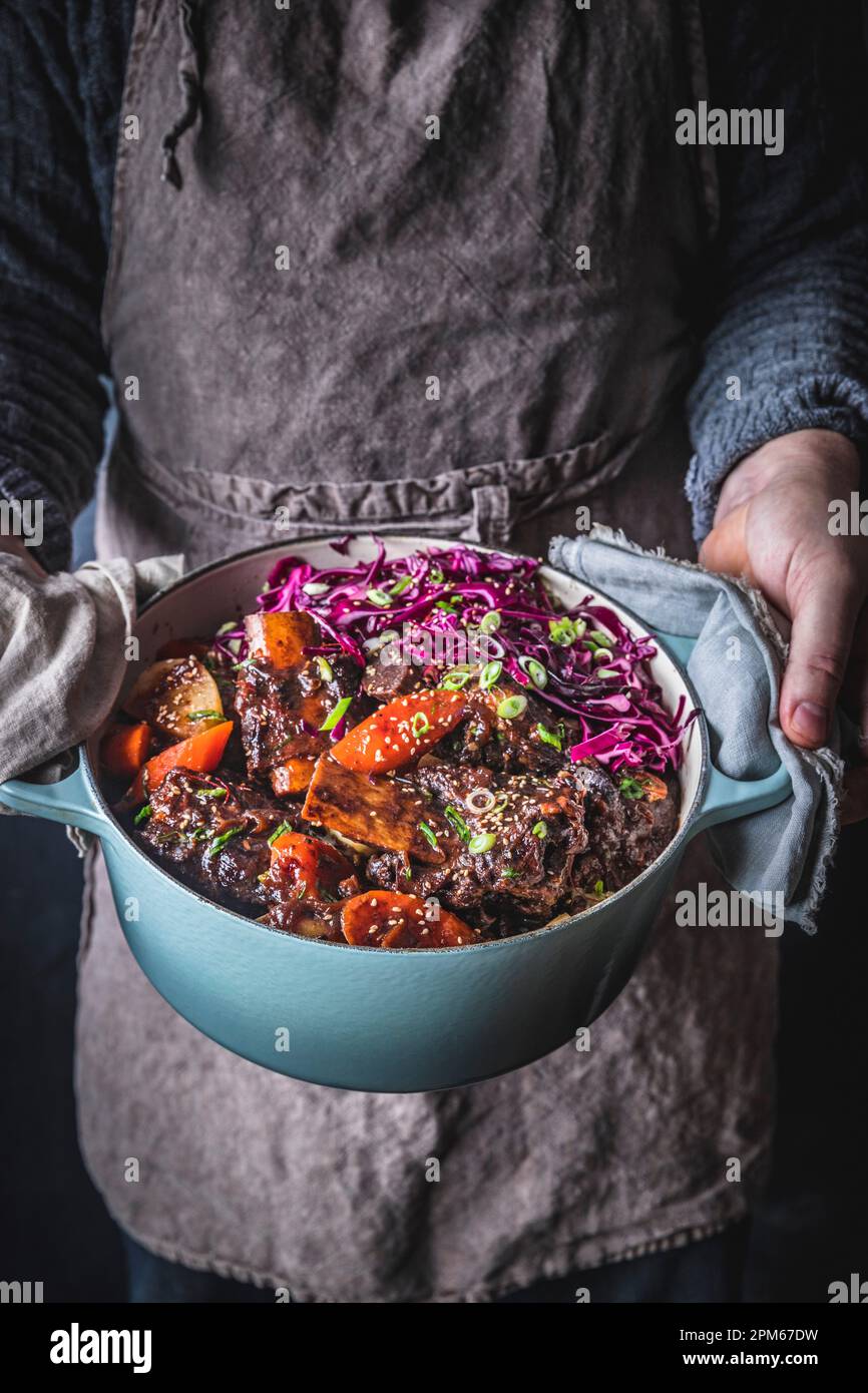 Man in gray apron holding Dutch Oven with Braised Short Ribs, carrots ...