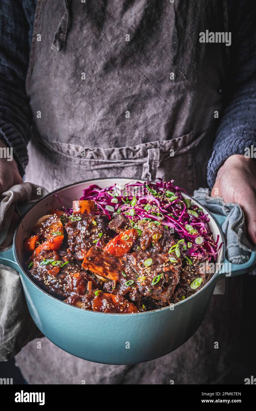 Man in gray apron holding Dutch Oven with Braised Short Ribs, carrots ...
