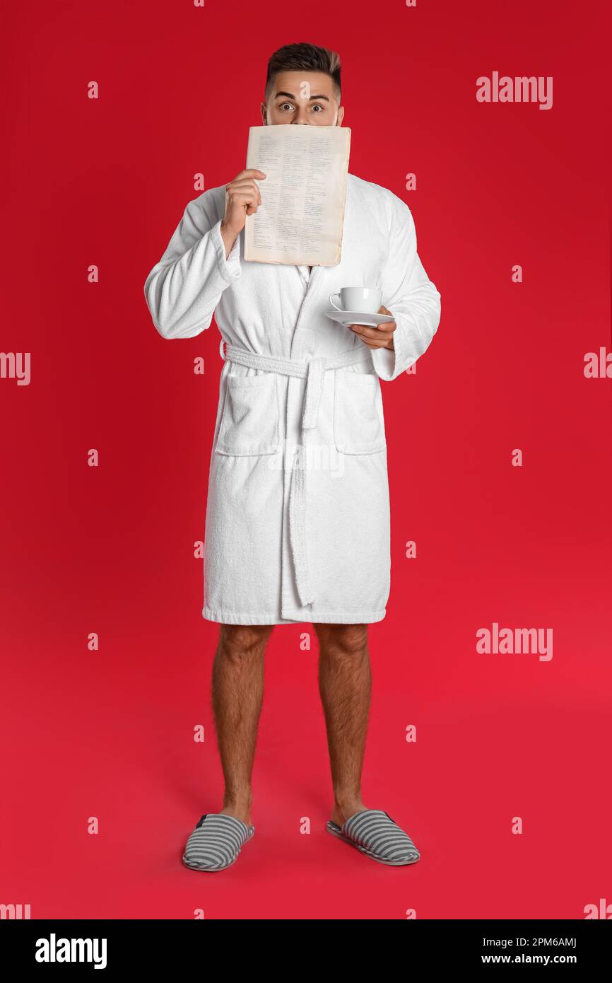 Young man in bathrobe with cup of coffee reading newspaper on red background Stock Photo