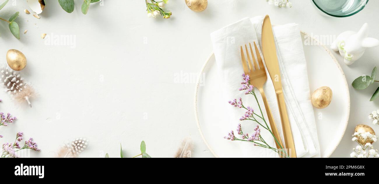Easter table decorations. Stylish Easter brunch table setting with white plate, napkin, golden cutlery, easter eggs and spring branches on light grey Stock Photo