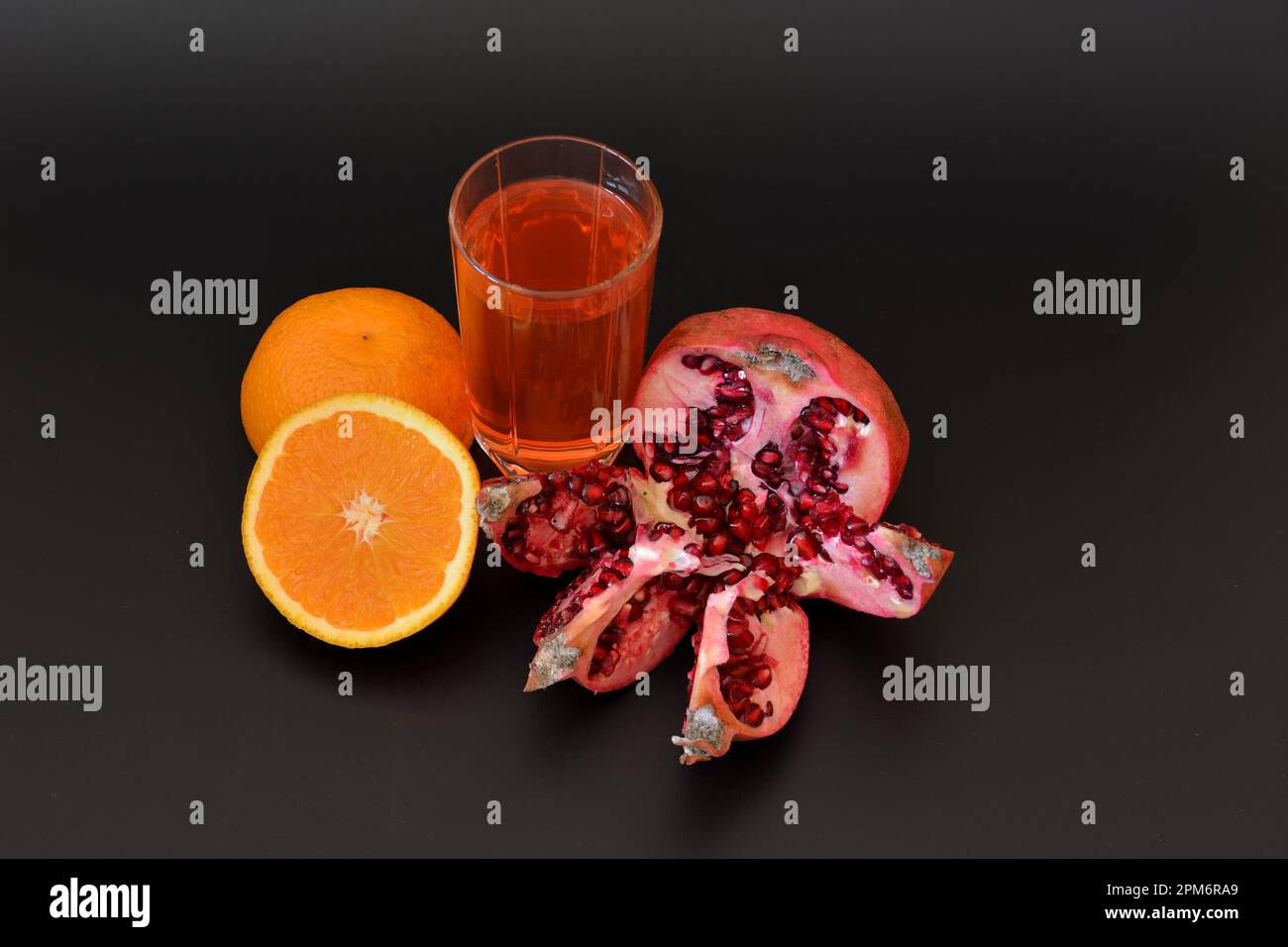 A transparent glass cup of a mixture of freshly squeezed tropical fruits on a black background, next to it is a half of a ripe orange and a broken pom Stock Photo
