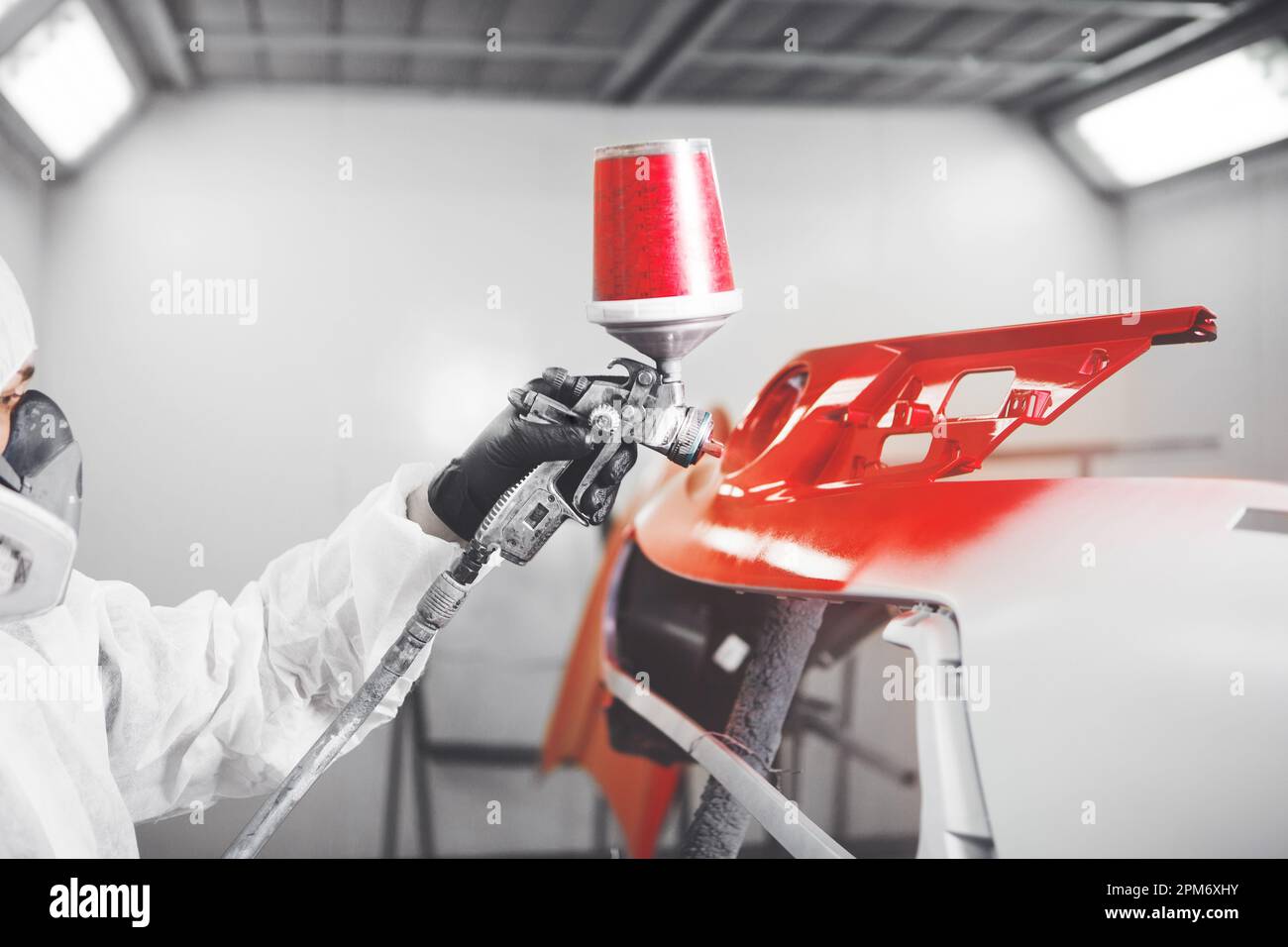 Spray painter worker in protective glove with airbrush pulverizer painting red car bumper in white paint chamber. Stock Photo