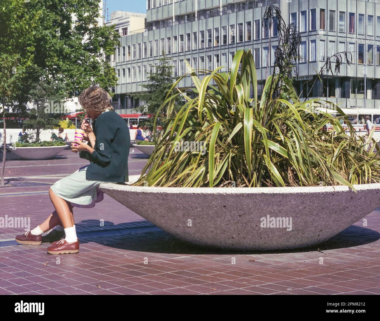 A 1981 historic image of two teenagers in Christchurch Girls High School (CGHS) uniforms sitting on the edge of a large dish potplant in front of the BNZ building in Cathedral Square, Christchurch, on the South Island of New Zealand Stock Photo