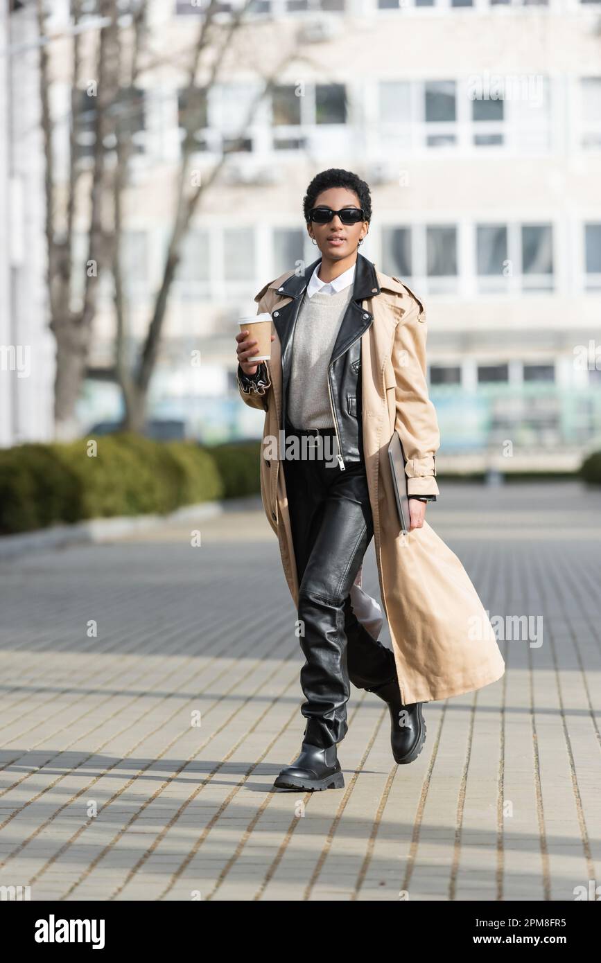 full length african american businesswoman in black leather pants and trench coat holding laptop and paper cup while walking on blurred street,stoc Stock Photo - Alamy