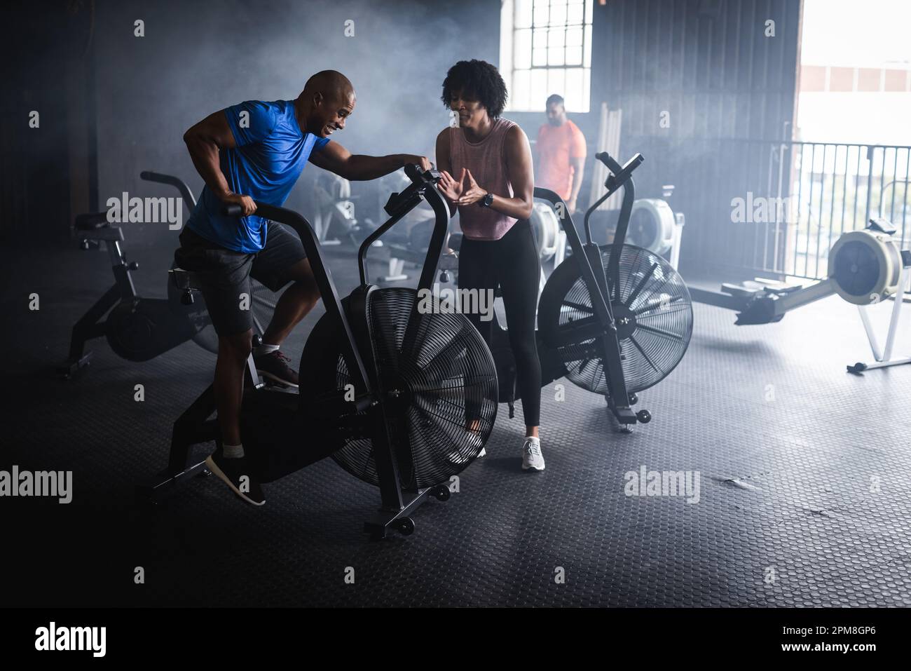 Biracial young woman clapping hands and cheering male friend cycling on exercise bike in gym Stock Photo