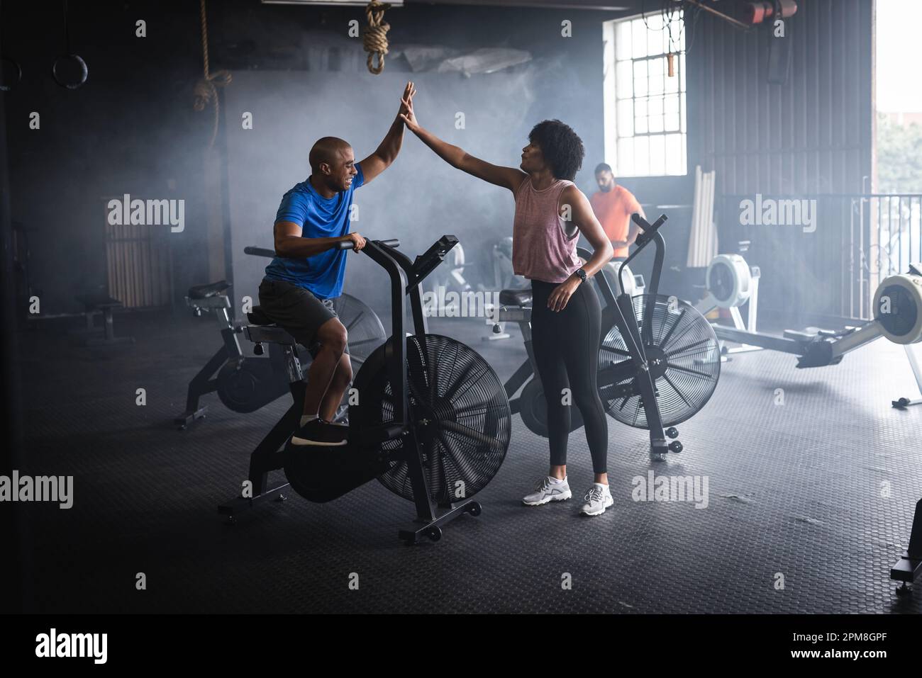 Biracial young woman giving high five to male friend cycling on exercise bike in gym, copy space Stock Photo