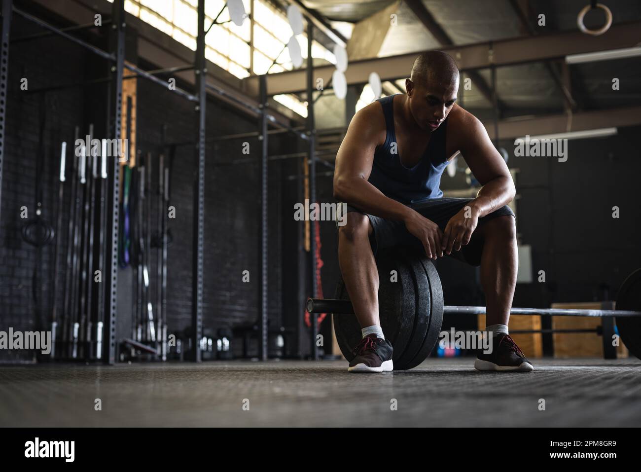 Full length of tired biracial young man with shaved head sitting on barbell in gym, copy space Stock Photo