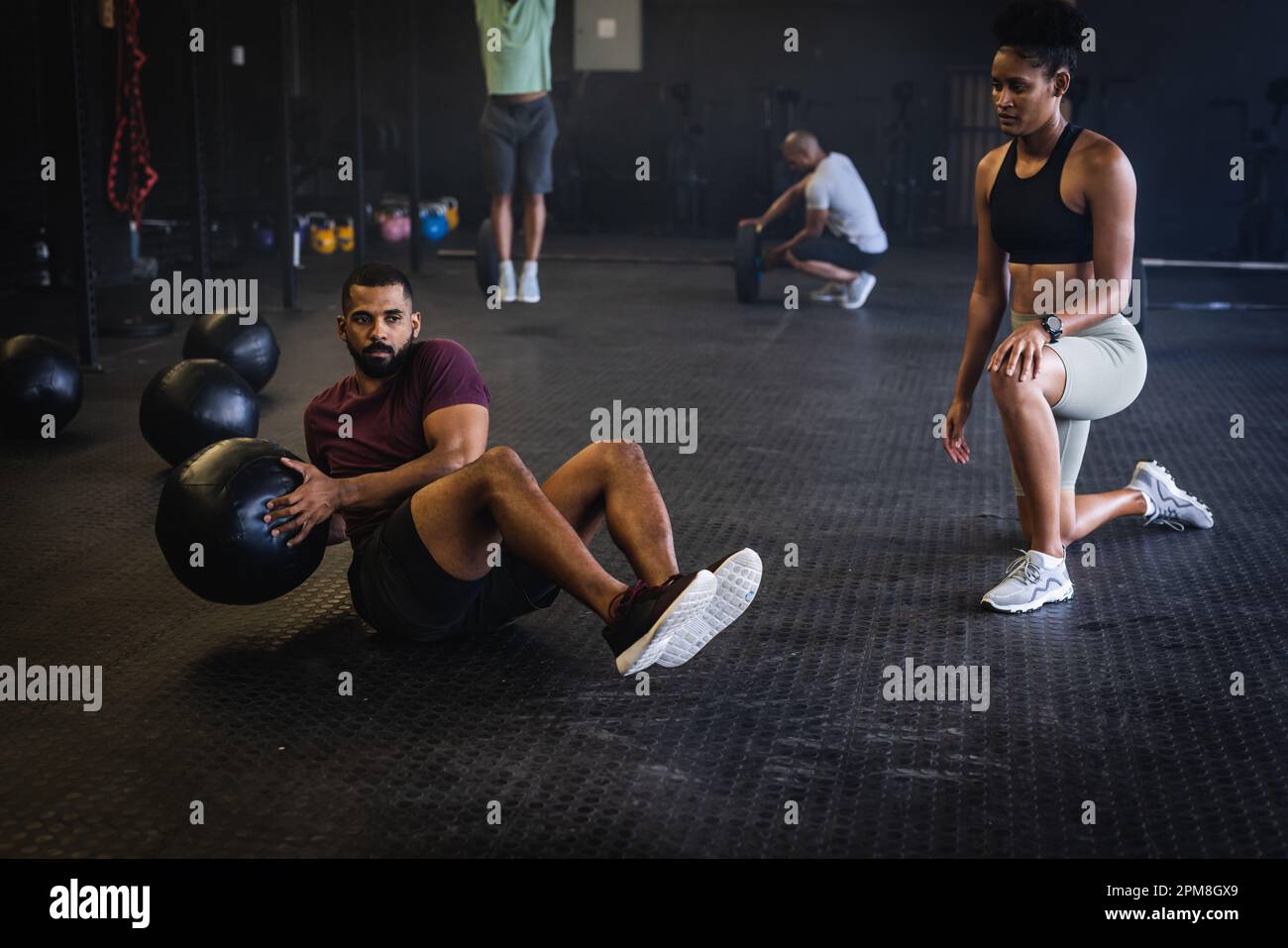 Biracial young woman looking at male friend exercising with fitness ball while lying on floor in gym Stock Photo