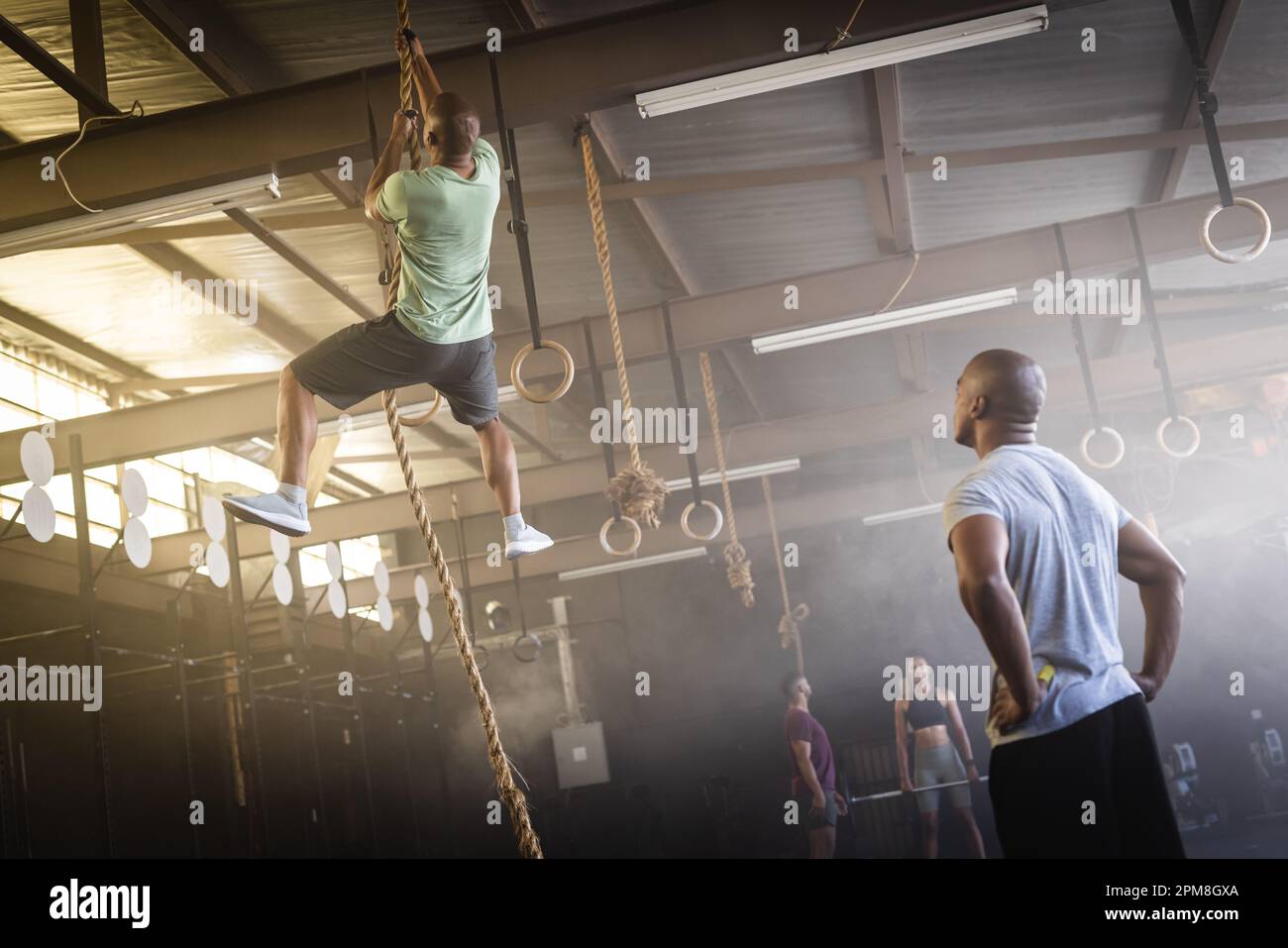 Biracial young man looking at male friend climbing on rope in gym, copy space Stock Photo