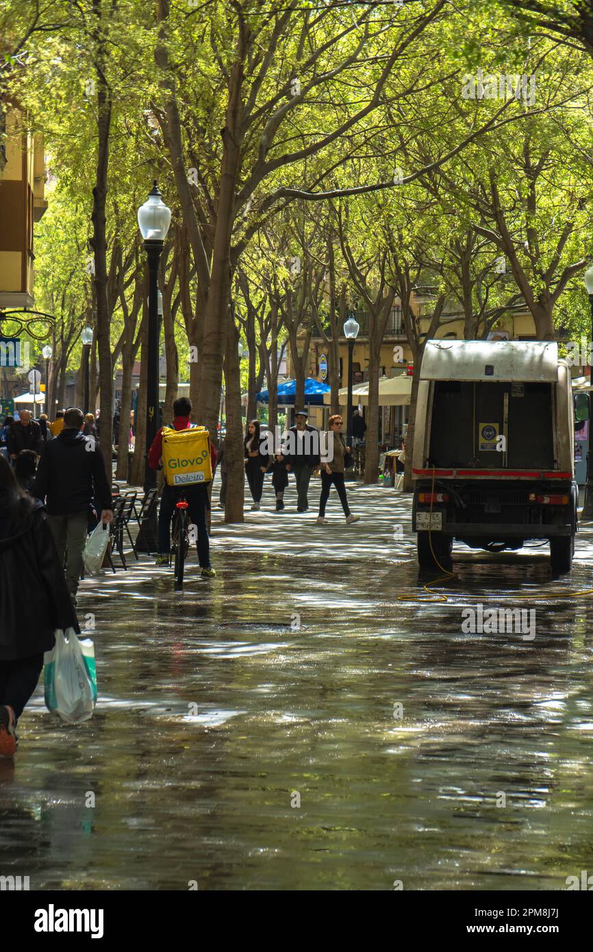 Barcelona municipal service truck, cleaning a pedestrian street with green trees and delivery man on a bicycle and pedestrians walking on the wet grou Stock Photo