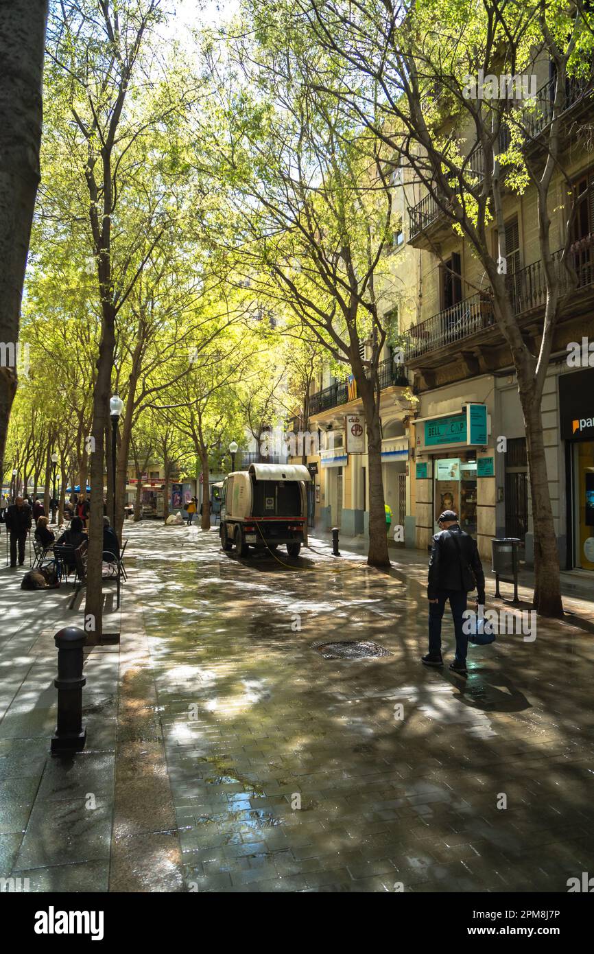 Truck of the Barcelona municipal service brigade, cleaning a pedestrian street with green trees and pedestrians walking on the wet ground Stock Photo