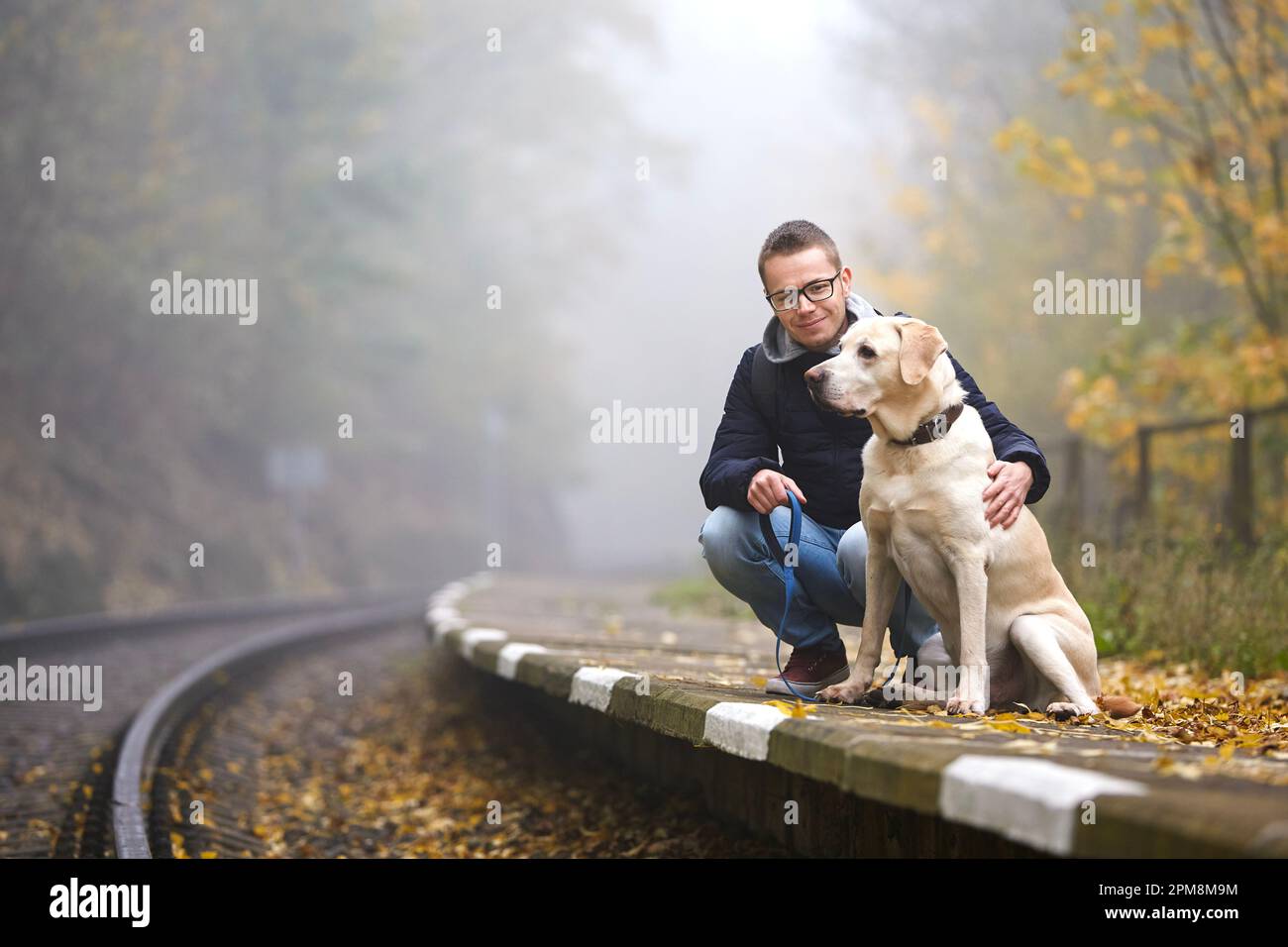 Man with dog on leash are waiting on platform of railway station. Pet owner and his labrador retriever together during trip by train. Stock Photo