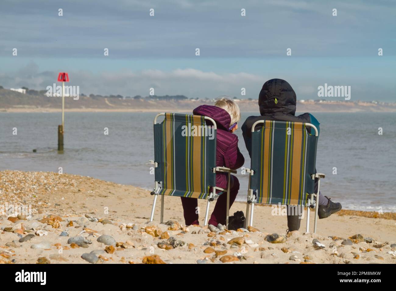 Two People In Overcoats Wrapped Up Against The Cold Sitting On Foldaway Chairs Enjoying The Winter Sunshine, Avon Beach, Christchurch UK Stock Photo